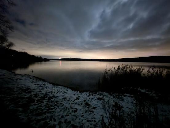 Written with the help of ChatGPT:

This photo shows another perspective of the Havel. The shallow, slightly snow-covered bank can be seen in the foreground, gently merging into the calm water. The surface of the water is smooth and reflects a vertical, reddish light, presumably from a signal or mast on the opposite bank. The dark outlines of trees and a single structure, possibly a tower or mast, can be seen on the horizon. The sky is covered by dense clouds, but shows a slight brightening in the distance, presumably due to light pollution from urban areas. The scenery appears calm and restrained, with a slight melancholy.