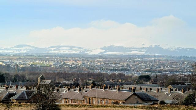 Rows of red brick terraced houses with slate roofs and a church can be seen in the foreground, followed by bands of forest, new builds and other houses before reaching the foothills of the Pentlands hills which have patches of frost covered fields, dark forest and cliffs but still capped by snow and ice which fades into hazy white clouds and a pale blue sky.