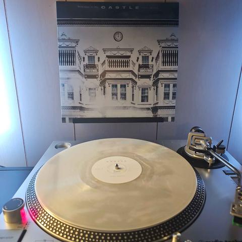 A Sunrise Pearl vinyl record sits on a turntable. Behind the turntable, a vinyl album outer sleeve is displayed. The front cover shows the front of a large manor/mansion.
