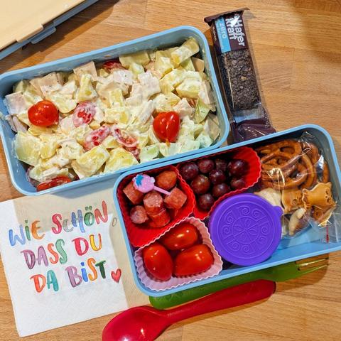 Holztisch, darauf 2 Ebenen einer himmelblauen Bentobox aus Plastik. 1 Ebene ist gefüllt mit Crackern, Weintrauben, Salami-Stückchen in denen ein Spießchen in Erdbeerform steckt, Mini-Tomaten und einem Döschen mit Dip. Die 2. Ebene mit Kartoffelsalat und einem weißen, cremigem Dressing. Daneben liegt eine Serviette mit der bunten Aufschrift "Schön, dass du da bist!", wiederverwendbare Plastikgabel und Löffel und ein Zero Zucker Schoko-Haferriegel.