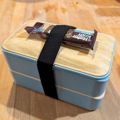 Holztisch. Darauf eine geschlossene Plastikbentobox mit 2 Ebenen. Sie wird von einem breiten Gummi zusammengehalten. Obendrauf klemmt noch ein Schoko-Haferriegel dazwischen.