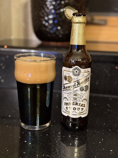A point glass of dark beer with a thick tan head. The brown bottle has a gold foil neck label holding the bottle cap and a white body label with gold and black lettering