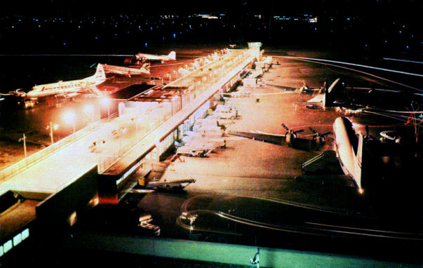 The view is at night, with both sides of the terminal light with very orange lights. You see prop planes on both side. On the right are two United Airlines DC-6 airplanes. On the left, a Captial Airlines Viscount, and two unidentified DC-3 planes.
