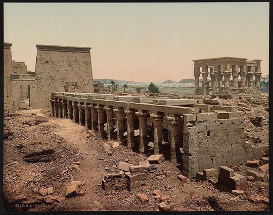 This image depicts an archaeological site with ancient Egyptian structures. The scene shows a row of tall, cylindrical columns forming what appears to be the remains of a colonnade or entranceway leading into larger temple-like buildings in the background. These large edifices have intricate hieroglyphic carvings on their walls and are constructed from stone blocks. Some pillars still stand intact while others lie broken around them. The site is surrounded by dry, dusty ground with scattered bricks and stones indicating excavation or decay over time.

In the distance beyond these ruins, palm trees and a serene landscape can be seen under what appears to be a hazy sky, suggesting an arid environment possibly indicative of desert surroundings typical for ancient Egyptian sites like Philae. The overall impression is one of historical grandeur now in states various degrees of preservation or ruin. This particular print was likely produced by the Photoglob Company and captured during roughly 1890-1910 period, reflecting early photographic techniques used to preserve images from such archaeological explorations at that time.