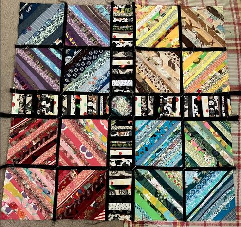 A square string quilt in progress, with the quadrants of blue/purple, yellow/tan, red/pink, and blue/turquoise strips, separated by narrower panels of black/white strips; all separated by narrow black strips. It is pinned in several places with safety pins.