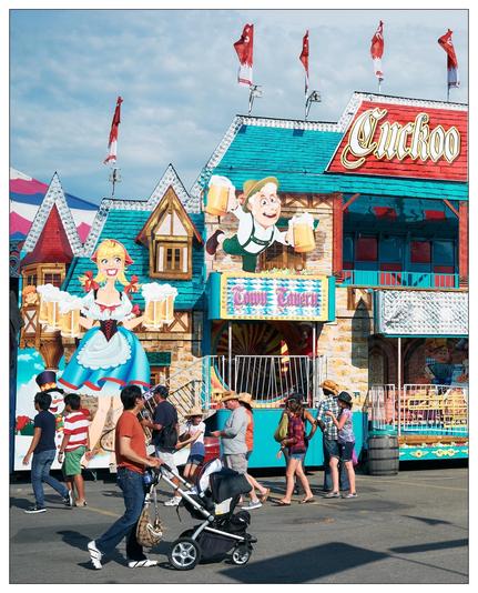 This image shows a lively street scene at a fair or festival. The building in the background, labeled "Cuckoo", is designed with a colorful, Alpine theme, reminiscent of a German beer hall. It features two large, cartoonish figures—a male character holding beer steins and a female character dressed in typical Bavarian attire. The architecture includes pointed roofs typical of European cottages, adding to the festive and thematic ambiance.

In the foreground, various people are walking and interacting. There's a man pushing a baby stroller, and several other individuals who appear to be enjoying the day out, some in casual outfits and others dressed in more distinctive costumes, such as the person resembling "Where's Waldo". The environment is lively, with bright colors enhancing the festive feel of the scene. Flags and additional decorative elements suggest a themed event or section of the fair dedicated to celebrating a specific cultural heritage, possibly a German beer festival based on the visual cues.