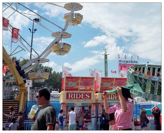 This image depicts a lively scene at a fair or amusement park. Featured prominently is a Ferris wheel with yellow and white gondolas. In the foreground, various people are seen enjoying the park, including a man drinking from a large cup and a woman looking up, possibly shielding her eyes from the sun or watching something. There are also several vendor booths advertising tickets for rides, snacks, and other attractions. The environment is colorful, with multiple flags and banners flying in the background, adding to the festive atmosphere of the scene.