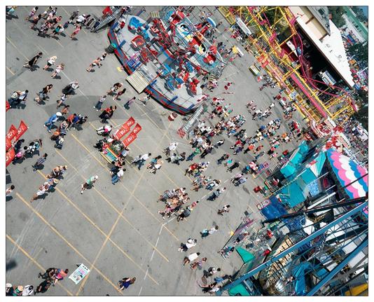 This image captures an aerial view of a bustling outdoor fair or amusement park. The scene includes a variety of colorful amusement rides, such as a ferris wheel and roller coasters. You can see many people walking around or gathering in groups throughout the park, indicating a lively atmosphere. The photo conveys a sense of activity and enjoyment, typical of a fair environment.