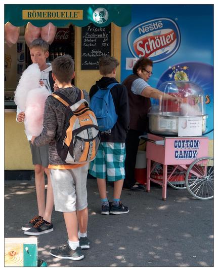 In the image, you see a group of three boys at a cotton candy stall. The boys are holding a large cotton candy each.  The background features signage that includes the brands “Coca-Cola” and “Nestle Schöller”. It appears to be a sunny day and the setting seems to be casual, perhaps at a fair or outdoor market. The atmosphere is lively and colorful, suggestive of a leisurely, enjoyable day out.