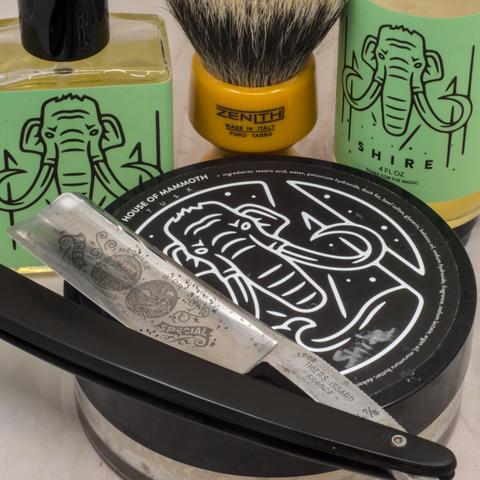 (ChatGPT description)

This image showcases a traditional wet shaving setup, featuring several artisan and vintage items. The centerpiece is a straight razor with a visible patina on its blade, engraved with intricate designs and text, indicating a vintage origin. The razor rests on a tub of shaving soap, branded "House of Mammoth," with a distinctive logo of a mammoth head against a black background.

To the right, there's a bottle labeled "Shire" with a green background and matching mammoth logo, likely a complementary aftershave or shaving product. A yellow shaving brush with a "Zenith Made in Italy" stamp on the handle sits in the background, hinting at its craftsmanship and high-quality natural bristles. Completing the setup, a clear square bottle with the same green mammoth branding is placed on the left, likely cologne or another shaving accessory.

The overall arrangement emphasizes a theme of elegance and tradition, appealing to enthusiasts of wet shaving and classic grooming rituals. The mammoth logo adds a contemporary and artisan touch to the vintage aesthetic.