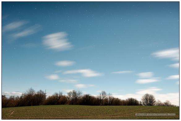 Für alle Fotos: Kahle, leicht hügelige, winterliche Feldlandschaft im Licht des Vollmonds an einem klaren, mit wenigen Schönwetterwolken betupften Himmel, weitwinklig fotografiert, Teils sind Winkraftanlagen im Bild, deren Rotoren wegen der Bewegungsunschärfe annähern unsichtbar bleiben. Wegen der langen Belichtungszeiten erscheinen auch die Wolken ziehend-unscharf. Erstaunlich finde ich das Spektrum des Lichts des Vollmonds: Die Farben sind denen der Landschaft im Tageslicht sehr ähnlich, der Himmel erscheint blau und hell, wenn auch sternengesprenkelt. Dennoch bleibt ein schwer in Worte zu kleidender Unterschied in der Farbgebung: Irgendetwas ist anders als im Sonnenlicht..