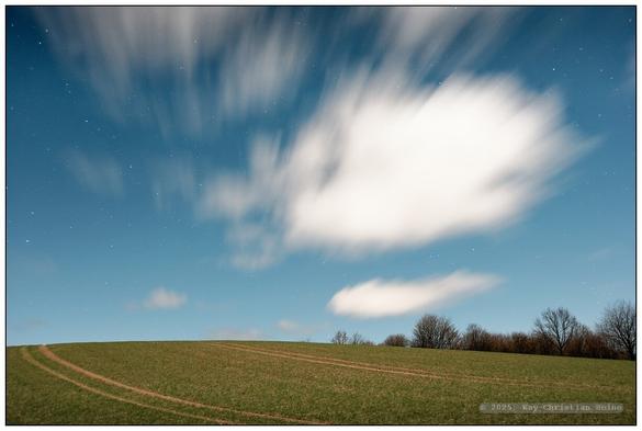 Für alle Fotos: Kahle, leicht hügelige, winterliche Feldlandschaft im Licht des Vollmonds an einem klaren, mit wenigen Schönwetterwolken betupften Himmel, weitwinklig fotografiert. Wegen der langen Belichtungszeiten erscheinen die Wolken ziehend-unscharf. Erstaunlich finde ich das Spektrum des Lichts des Vollmonds: Die Farben sind denen der Landschaft im Tageslicht sehr ähnlich, der Himmel erscheint blau und hell, wenn auch sternengesprenkelt. Dennoch bleibt ein schwer in Worte zu kleidender Unterschied in der Farbgebung: Irgendetwas ist anders als im Sonnenlicht..