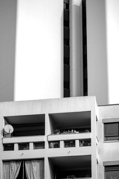 Photo noir et blanc d'un petit immeuble en béton, vu côté balcon, avec une antenne parabolique et quelques fleurs en pot.
En arrière-plan, un immeuble gigantesque et presque aveugle encombre toute la vue.