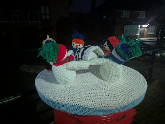 It is night time. Three snowmen and two penguins, all in woolly hats and scarves stand in a circle holding hands around an object which might be an igloo.
(they are knitted figures atop a postbox)