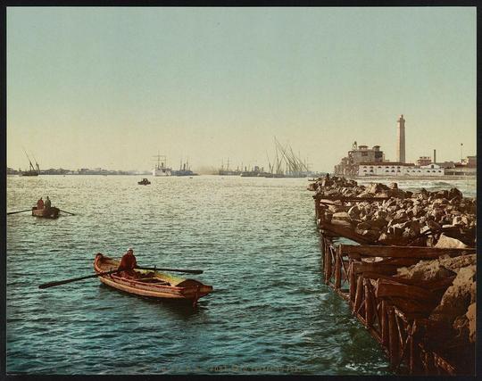 The image depicts a coastal scene with various elements that suggest it is from the late 19th or early 20th century, based on its style and subject matter. The foreground features two boats; one appears to be an old-fashioned rowboat with visible occupants wearing traditional clothing, while another boat is moored near a wooden jetty extending into calm waters. Beyond the waterway lies a bustling harbor filled with numerous ships of different sizes, indicating active maritime trade or military presence.

A prominent lighthouse stands on what seems like rocky terrain in the background, possibly serving as an aid to navigation for vessels approaching Port Said, which is known historically as one of many entrance points into the Suez Canal. The surrounding landscape includes a small industrial structure and buildings that may be warehouses or other commercial establishments associated with port activities.

The overall color tone has hues reminiscent of sepia, suggesting either age or artistic stylization to evoke an antiquated feel. Notable features include calm waters reflecting sunlight, creating a serene atmosphere amidst the busy harbor backdrop. The composition effectively captures both the tranquil and industrious aspects characteristic of Port Said's maritime history during that period.