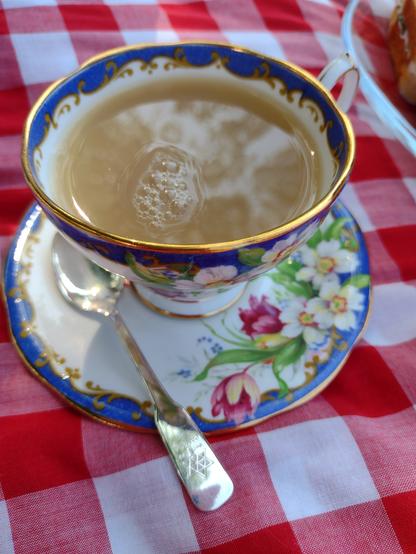 Close up of a cup of tea in a China teacup. The tea in the cup has a few bubbles in it and a reflection of the trees above the picnic are visible. The cup has a blue and gold (as in metallic gold) decorated rim on a white background. The saucer has pink and yellow tulips and a yellow English rose and the blue and gold scrolled pattern. A 1967 silver teaspoon commemorating Canada's 100th Birthday rests in the saucer. The cup and teaspoon sit on classic red and white gingham tablecloth (placed on the grass). A glimpse of a sandwich can be seen in the upper right corner.