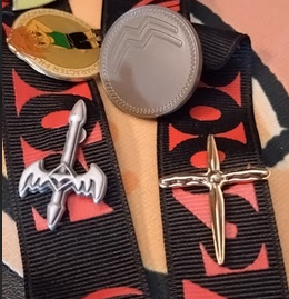 Close up of three enamel pins, the top is a round bronze coin with textured edge and two double rising points at the top. The others are a pair of swords in embossed metal, one is silver with a sword with bat winged hilt and diamond shaped center. The other is gold, with a cross like shape and slightly waved hilt ends.