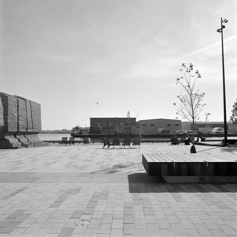 Clean paving bricks stretch to the end of the Western Gap with the Island Airport service buildings on the far side. Deck chairs in groups, a scant tree, wooden benches, and the grey stone of the Irish Famine memorial to the side as a seagull wings overhead.