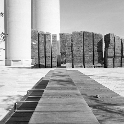 A wooden bench stretches toward a gap in the Irish Famine memorial, between the grey stone edifices a glimpse of the parkette beyond can be caught as the pale concrete of the malting towers rise to the left.