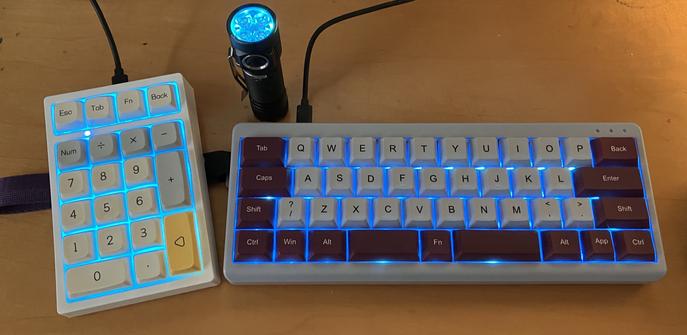 The image features a mechanical keyboard on the right with cyan backlighting and a numpad on the left, also illuminated in cyan. A small flashlight is positioned nearby with a cyan standby light. The scene is set on a wooden surface.