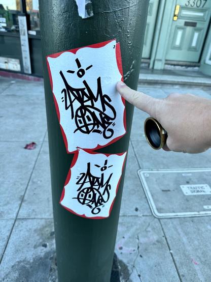 Two large stickers on a light post. Both stickers look like name tags that you'd write your name on, but these are comically large, like the size of a a 8.5" by 11" sheet of paper. Graffiti artists use name tags like this to draw their names on (aka "Sticker Slaps"), and these have been graff writing on them that appears to say "2 Atoms One" in a cool handwritting style.