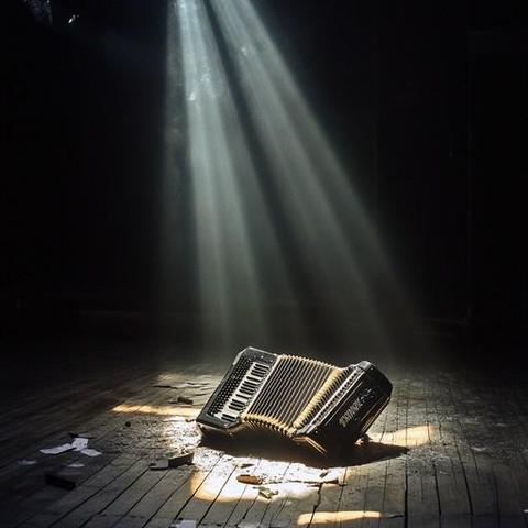 An AI-generated piano accordion sits, smashed, as though fallen from a great height, on a rubble-strewn stage floor... while sunlight filters through the dusty air through a hole in the ceiling (through which the instrument initially fell?) to shine down on it.