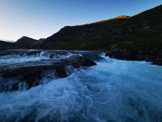 A rushing river flows over dark rocks, creating whitewater rapids. In the background, a range of dark, mountainous terrain is visible under a clear, twilight blue sky.