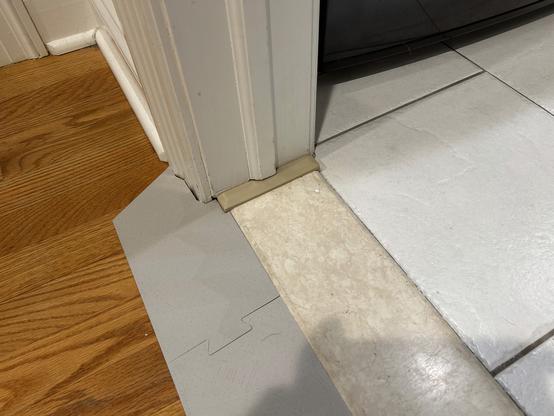 Photo of an indoor door sill.  To the left, the floor is hardwood.  To the right, it is tile.  In the middle is a marble-looking stone door sill, 134 millimeters wide and 20 millimeters high on the hardwood side.  A 3D printed ramp is on the hardwood side.  A 3D printed strap goes from under the ramp end, over the end of the door sill and down around the tile side of the sill.  It is tucked under part of the wood molding.