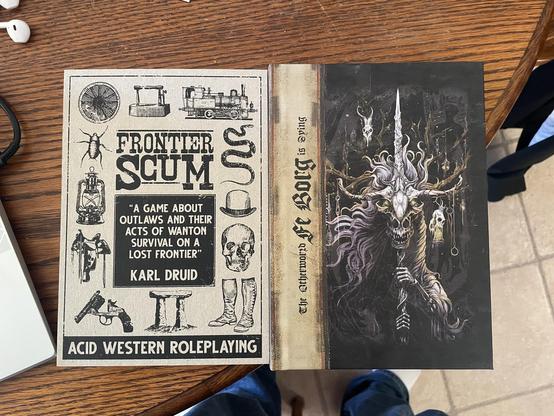 The image shows two tabletop roleplaying game books side by side on a wooden surface. The left book, titled "Frontier Scum," features a vintage design with illustrations of outlaws, firearms, and theatrical elements. The right book, titled "Fe Borg” features a strange and horrific illustration of a draconian skull and bone sword grasped by skeletal hands merging together.