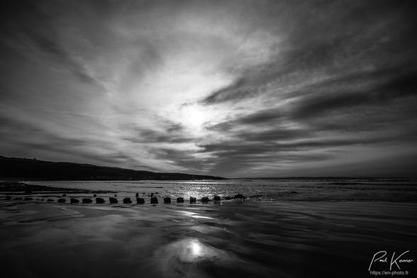 Photo monochrome en noir et blanc, présentant dans les deux tiers supérieurs, un halo solaire dans un ciel, partiellement nuageux et dans le tiers inférieur le haut des membrures de la carcasse d'un ancien bateau ensablé sur une grève. La marée montante arrive presque au niveau de la carcasse et le sable humide reflète le soleil et les nuages.