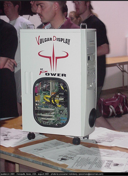 This image appears to show a custom-built computer case on display at a tech or gaming event. The case is branded with the text "Vulgar Display of Power," referencing Pantera's famous album. It features a bold, stylized Quake logo, suggesting a connection to gaming culture, possibly a Quake LAN event or convention.

The computer case itself is highly customized, with a large window on the side to showcase internal components. It is mounted on small wheels for portability, which is unusual for computer cases, and has a white, industrial design. The visible internals include neatly arranged hardware with color-coordinated cables.

Based on the event label, this was taken at QuakeCon 2001 in Mesquite, Texas, a popular annual convention for Quake and other PC gaming enthusiasts. The aesthetic and setup reflect the early 2000s modding and LAN party culture.