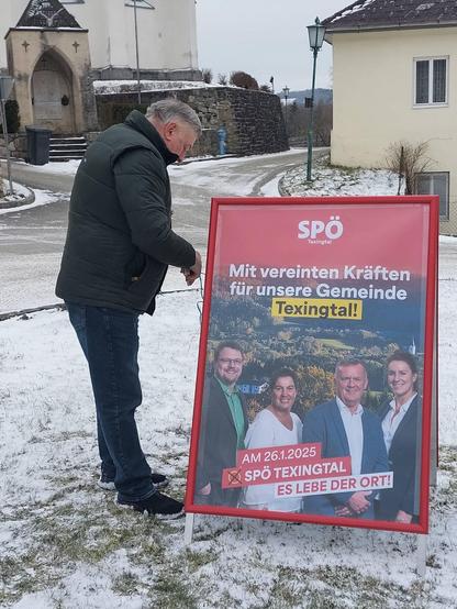 Ein Mann in Winterjacke befestigt einen Plakatständer mit einem Wahlplakat der SPÖ Texingtal. Auf dem Plakat steht der Satz Mit vereinten Kräften für unsere Gemeinde Texingtal!
Darunter sind die vier Kandidaten zu sehen, zwei Männer und zwei Frauen. 
Am Boden des Plakates steht der Satz Am 26.1.2025 X SPÖ Texingtal und der Slogan Es lebe der Ort!
