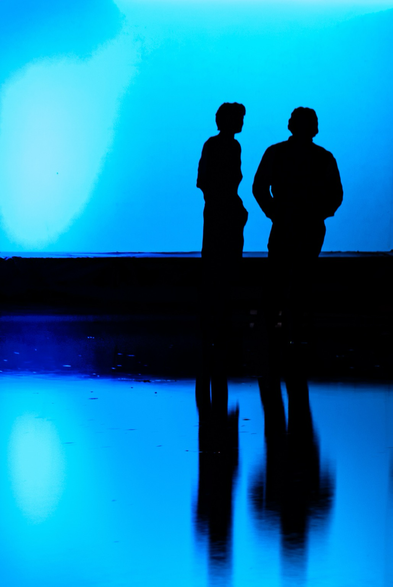Two silhouettes in front of a big blue screen. It seems that there are talking. Nobody knows.  The water is reflecting the whole picture.