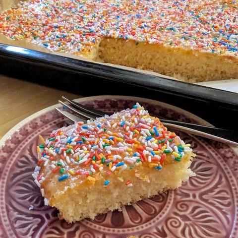 Kuchenteller mit einem Stück hellem Rührkuchen vom Blech. Das Stück ist glasiert mit Zuckerguss und mit vielen bunten Zuckerstreuseln bestreut.