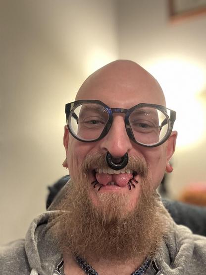 A bald person with a full goatee smiles at the camera, wearing large glasses and multiple facial piercings, including a black large gauge septum ring and six black lip rings. Their tongue is sticking out and split down the center and the background is softly illuminated.