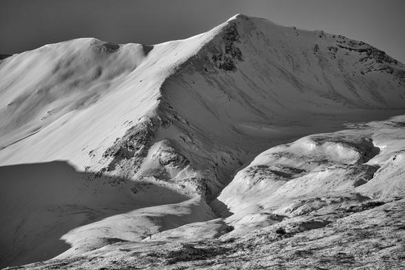 A black and white photo of a snow covered Beinn Eighe mountain. The sun cuts across from the top left creating shadows that contrast the snow well and bring out the ridgelines and valleys. Torridon, Wester Ross, Scottish Highlands.

Photo by and copyright of Paul Henni.