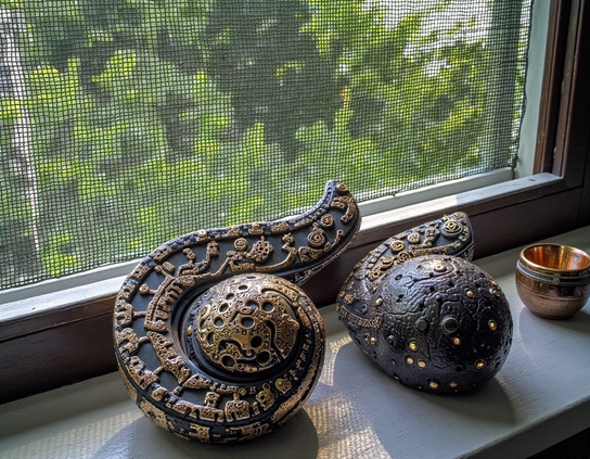 Two dried and intricately decorated black and gold gourds resting on a windowsill next to a small copper bowl, with sunlight filtering through the screen from a green outdoor view.