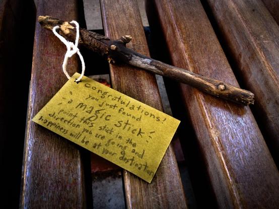 Overall Scene:
The image shows a wooden bench with a small, brown stick lying across it. A handwritten note is tied to the stick with a piece of string. The background appears to be a brick wall.

The Stick:
- The stick is a thin, brown branch with a few small knots and twigs.
- It looks like it was found in nature and is unprocessed.
- It is lying diagonally across the bench, with one end slightly off the edge.

The Note:
- The note is a rectangular piece of yellowish paper tied to the stick with a piece of white string.
- The writing on the note is in pencil.
- The writing says: "Congratulations! You just found a magic stick! Point this stick in the direction you are going and happiness will be at your destination."

The Bench:
- A CTA (Chicago Transit Authority) bench at the Western Brown line train station. 
- The bench is slightly weathered, with some visible wear and tear.

The Location: 
- The overall impression is one of whimsy and childhood imagination.