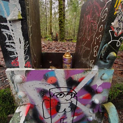 A metal structure, partially obscured by a graffiti-covered front panel, situated on a forest floor with leaves. The panel features a variety of vibrant colors, including pink, purple, and blue, with several abstract designs and text. A gold spray paint can with a purple label is positioned centrally within the opening of the structure, surrounded by colorful paint splatters, and a cartoonish drawing appears in black outline on the lower right section of the panel. The background shows a dense forest with tall trees and a layer of fallen leaves.