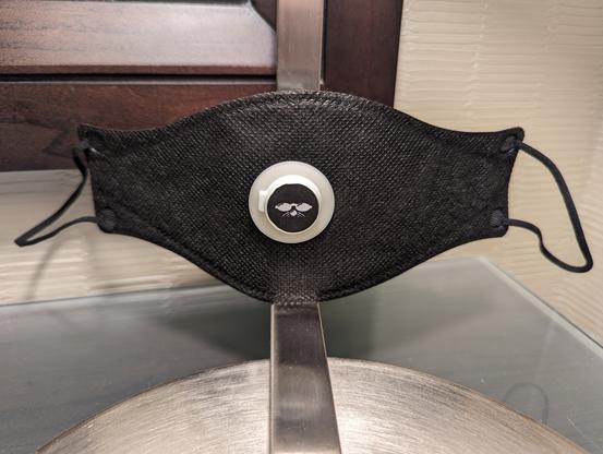Black CAN-99e respiratory mask with a SIPmask valve in the middle of the mask and a ShmooCon logo (black with white Shmoo face) on the valve cover.

(Mask is propped against the base of a lamp in the con hotel room.)