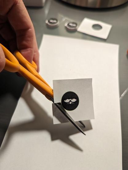 Cutting out a round ShmooCon logo from a square of paper using a small pair of orange scissors. There are two finished valve covers with ShmooCon logos in the background.