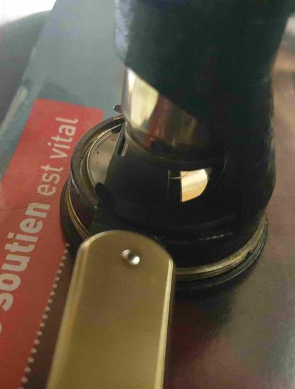 Un petit couteau pliant à manche laiton dont la lame en m390 est sous l'objectif d'un microscope miniature ancien à un oculaire un objectif plus zoom, le tout posé sur une lettre de médecins du monde sur laquelle on lit "votre soutien est vital".