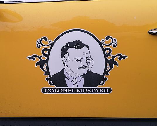 This image displays a black and white logo on the side door of a yellow Austin Mini. The main logo is slightly oblong, with scrollwork on both sides. In the center is a man with moustaches and a monocle,  wearing a suit.