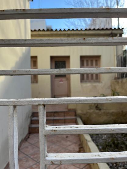 A metal gate of horizontal bars painted white in the foreground. Out of focus behind it, a blank wall on the left with a paved path leading back to a simple, small one storey house in the interwar vernacular style, tile-roofed and well-proportioned like a neoclassical house but unadorned and unassuming.