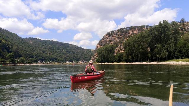 A person in a red canoe on the river. Further ahead on the right is the Loreley. Clothing and light appear summery. The sky is blue with a few clouds.