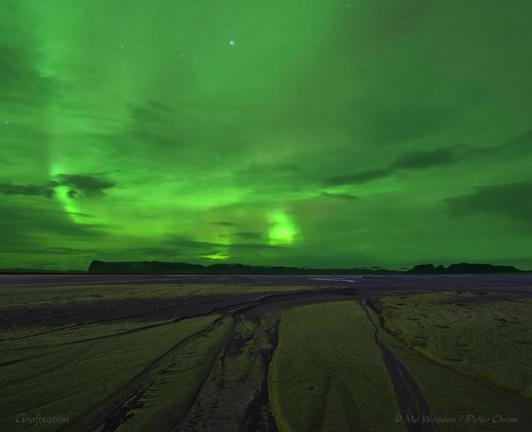A photo of a flat, sandy area of ground with water-cut channels running through it. It's late, and quite dark. On the horizon is the silhouette of a mountain range. The sky is patchy cloud and some stars are visible but bright green streaks of aurora are spread across the sky. This light is so bright that the sands below are tinged with colour.