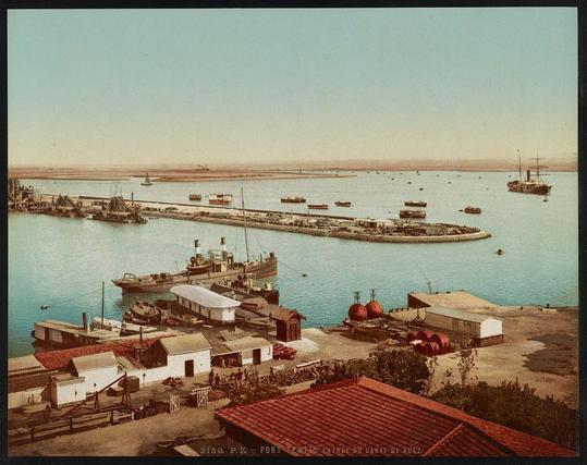 This image depicts a vibrant harbor scene with numerous boats anchored in the water. In the foreground, there's an industrial setting with buildings that have red roofs and various storage structures, including large cylindrical tanks possibly for fuel or chemicals. A prominent feature is a long pier extending into the sea where several ships are docked, indicating active maritime activity.

In the middle ground, the harbor continues to expand with more boats scattered in the water, suggesting significant commercial shipping operations. The background shows a wide expanse of calm blue waters leading towards distant landforms that appear as faint silhouettes against the sky. A small sailboat is visible on the horizon, adding a sense of scale and tranquility.

The overall color palette consists of muted earth tones with hints of red from the buildings' roofs and warm hues in some areas suggesting sunlight reflection or shadows. The composition captures both human-made infrastructure and natural elements harmoniously coexisting within this bustling harbor environment.