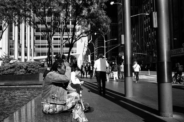 A black-and-white photo of a few people sitting on the edge of a fountain and some people walking away from the camera