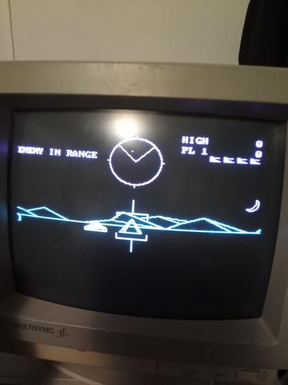 Battlezone game screen. Red radar and indicators, cyan terrain, and a white tank to shoot at.
