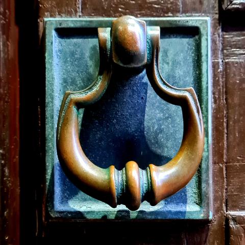 A curved brass door handle with some areas covered in a green patina, while others are polished and shiny.